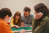 A family of two parents and two kids sit around a table with the Board console, holding playing pieces and interacting with the console